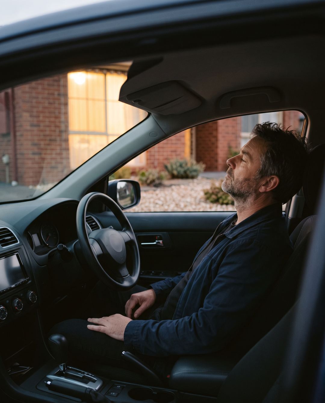 A man sitting in his parked car in the driveway at dusk, head back against the seat.